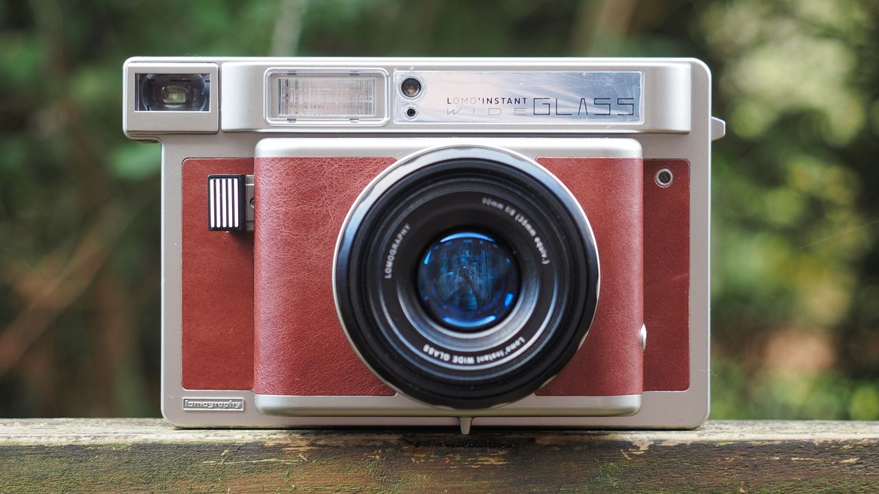 Lomo'Instant Wide Glass camera, sitting on a wooden surface, outdoors against a woodland background