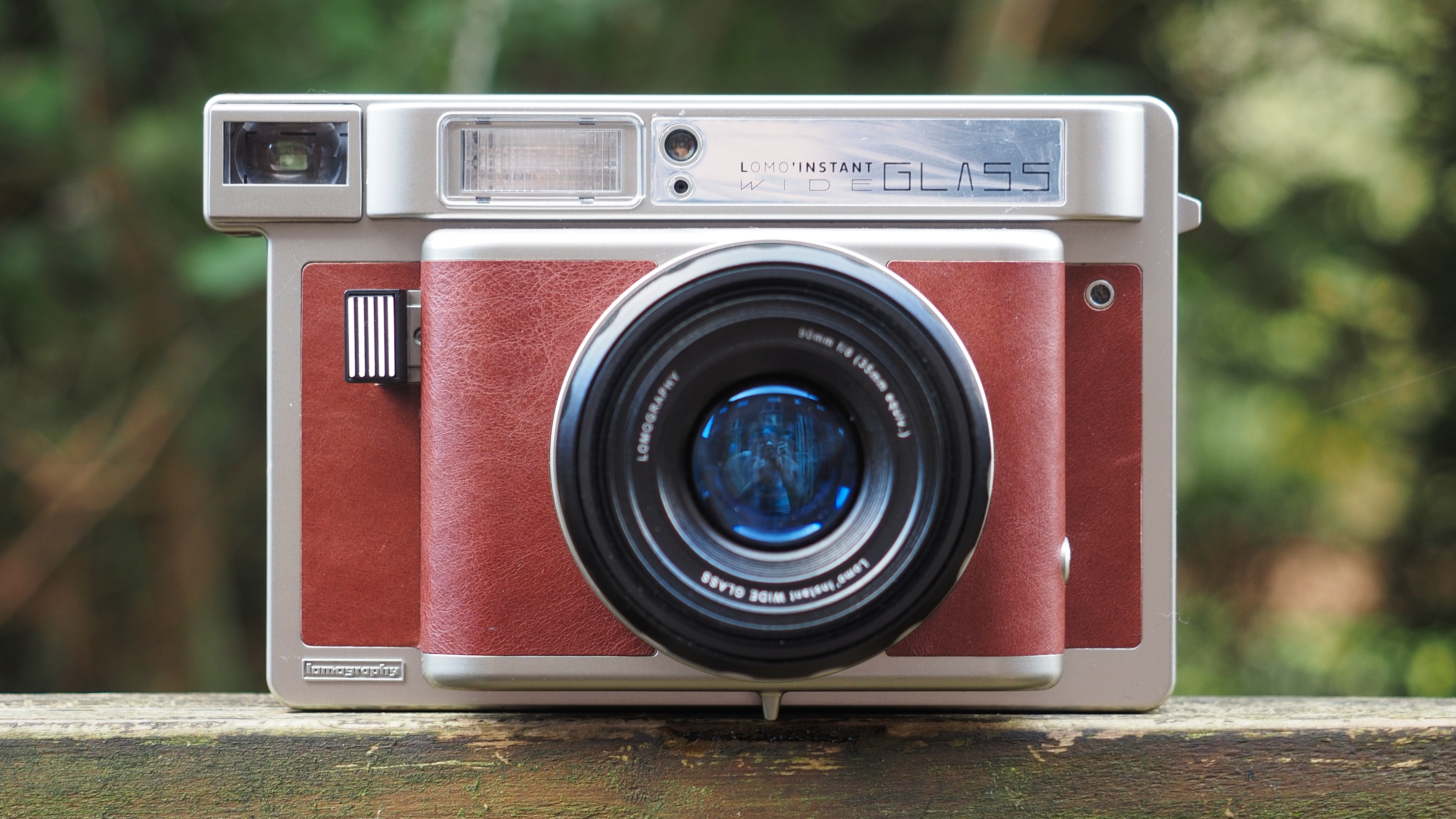 Lomo'Instant Wide Glass camera, sitting on a wooden surface, outdoors against a wooded background