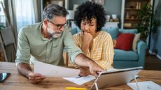 An older couple look over financial information on a laptop at their dining room table.