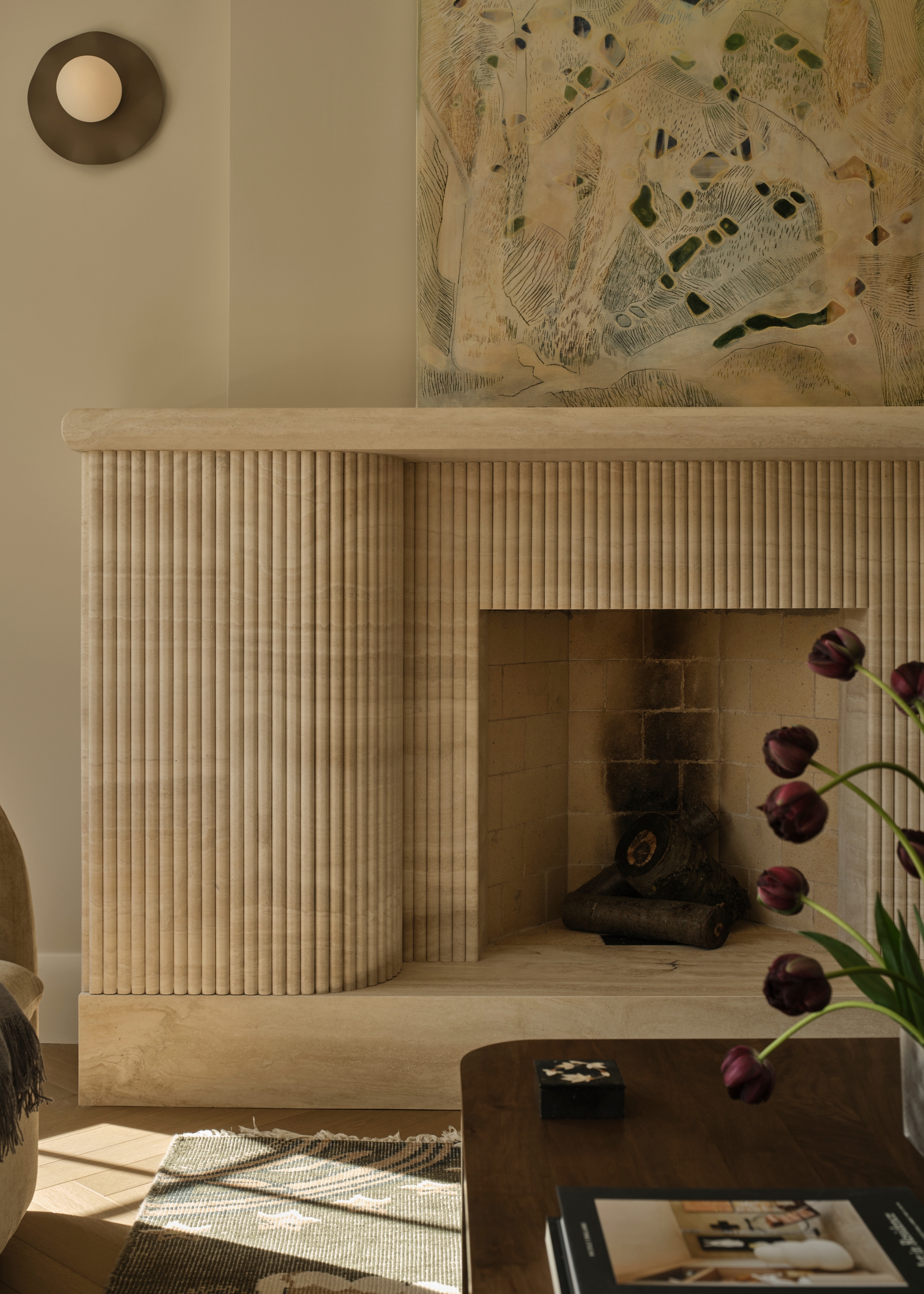 A living room with a ribbed stone fireplace, a leaning painting, a brass wall sconce, a vase of purple tulips, and a coffee-table book