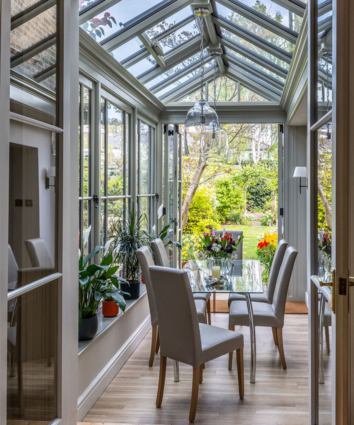 Inside a grey-framed orangery with a tall glazed pitched roof, a glass dining table with grey upholstered chairs, houseplants on the windowsill and open doors to a colourful spring garden.