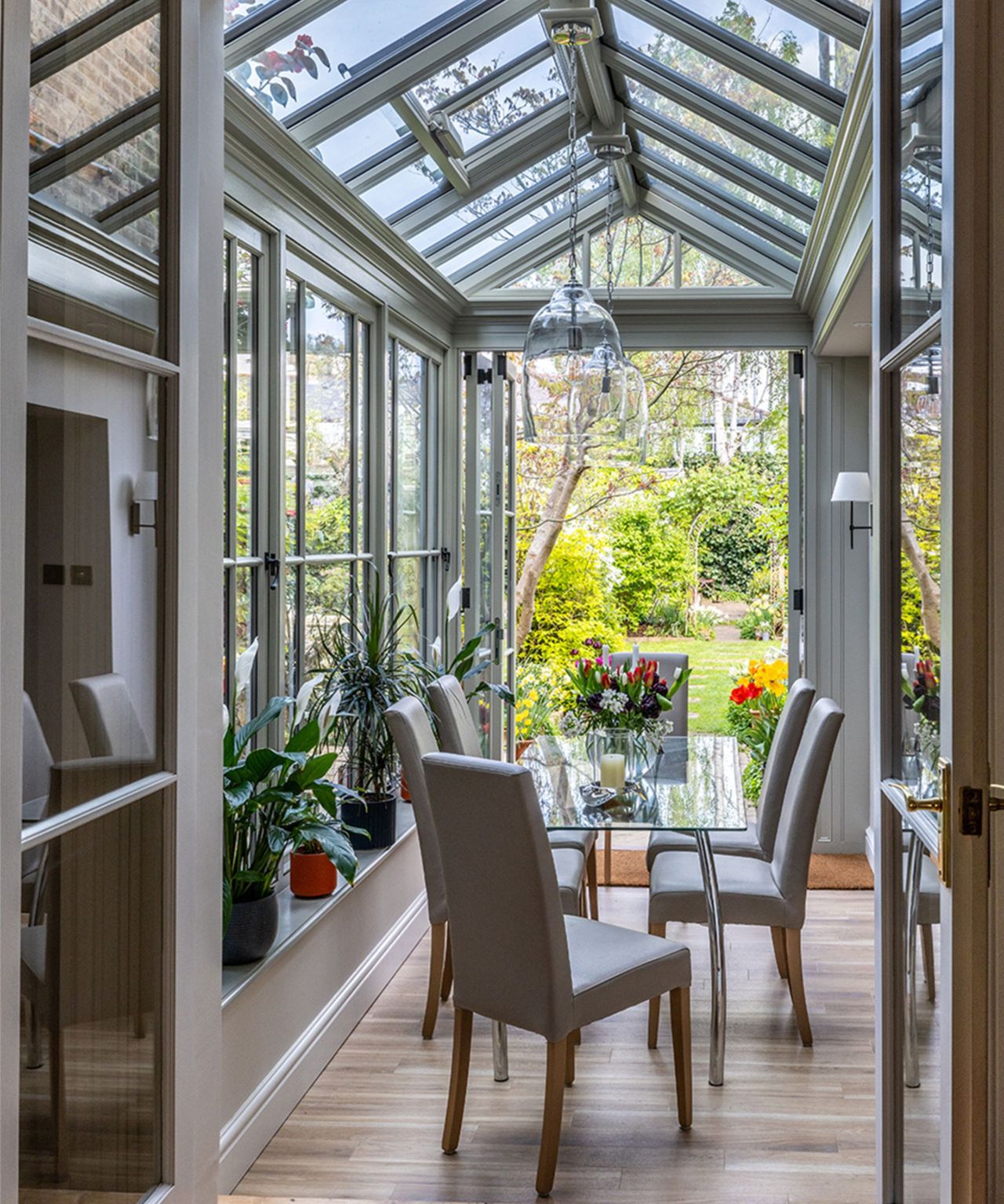 Inside a grey-framed orangery with a tall glazed pitched roof, a glass dining table with grey upholstered chairs, houseplants on the windowsill and open doors to a colourful spring garden.