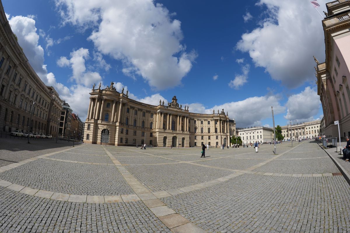 Ultra-wide view across a cobbled Berlin square lined with grand classical buildings beneath a bright blue and cloudy sky.