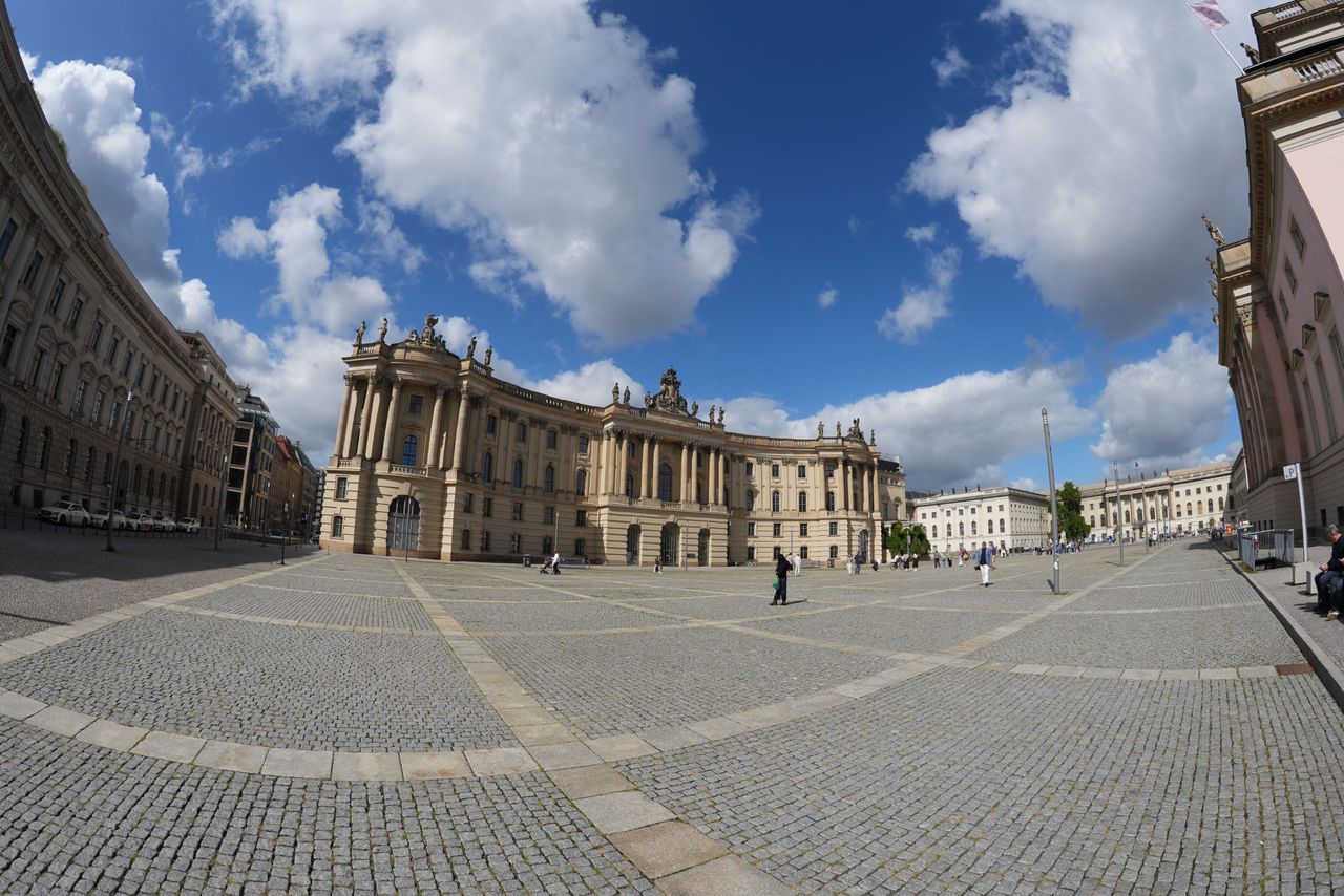 Ultra-wide view across a cobbled Berlin square lined with grand classical buildings beneath a bright blue and cloudy sky.