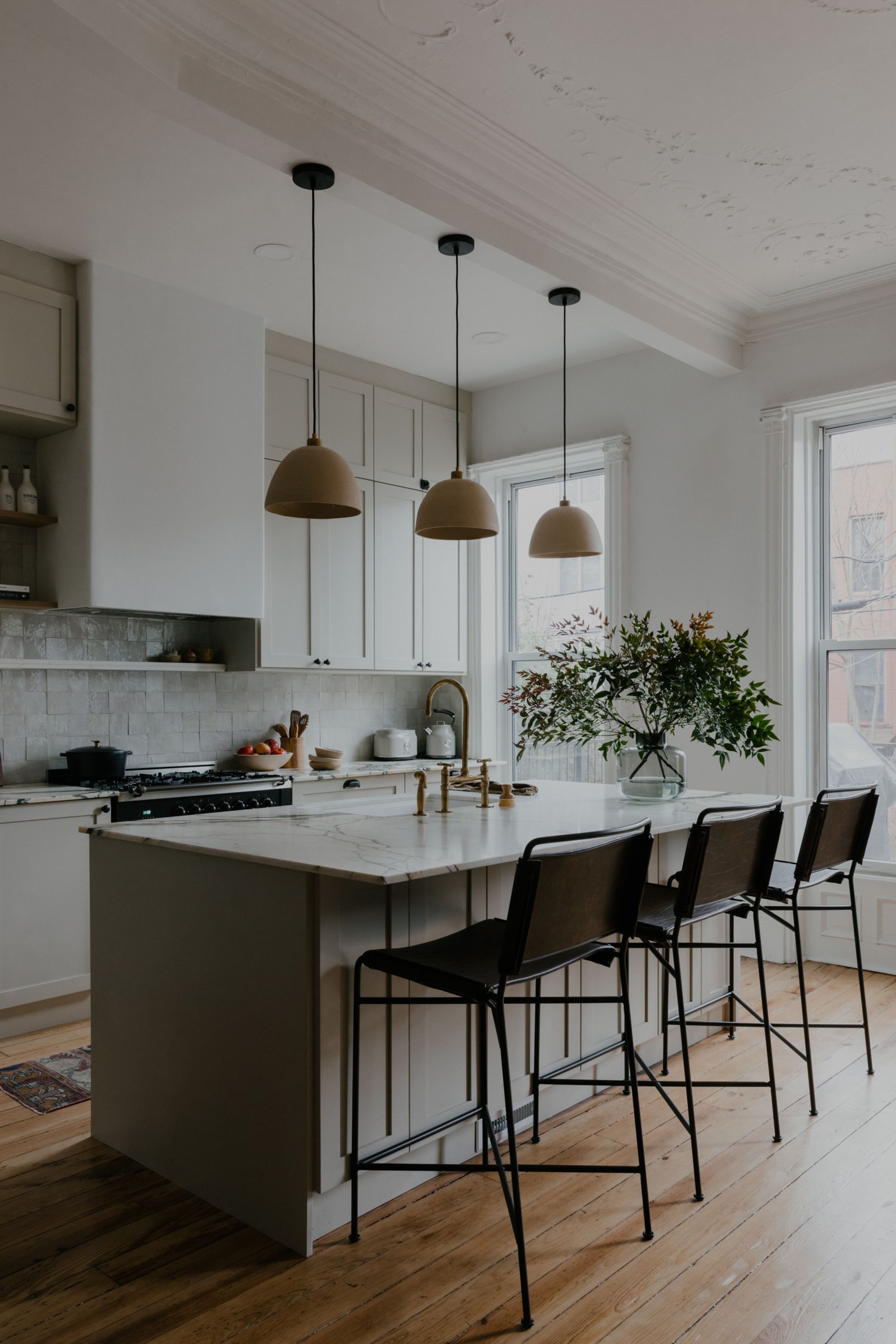 A modern neutral kitchen with three pendants over the island and three dark stools along one side with a large vase of foliage and flowers on top of the island