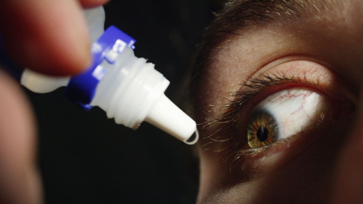 close up of a man&#039;s eye as he&#039;s squeezing a generic eye drop dropper near his face, pushing out a drop of clear liquid