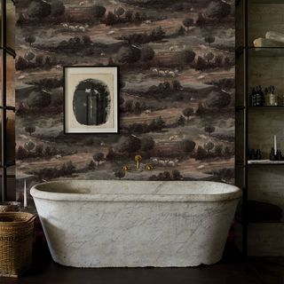 Image of a white marble bathtub in a dark room. There is dark green wallpaper with a pattern of trees and fields that looks like the English countryside