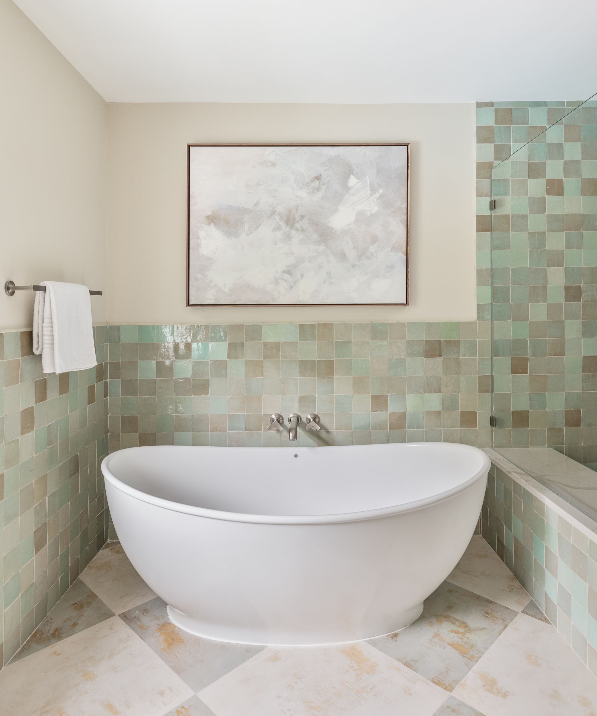 Minimalist bathroom featuring a white oval soaking tub against a wall of muted green and tan square tiles with an abstract painting above.