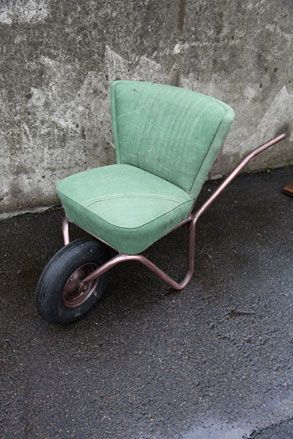 Concrete wall and floor, pastel green chair incorporated into a wheel barro, bronze frame and handle, black front wheel
