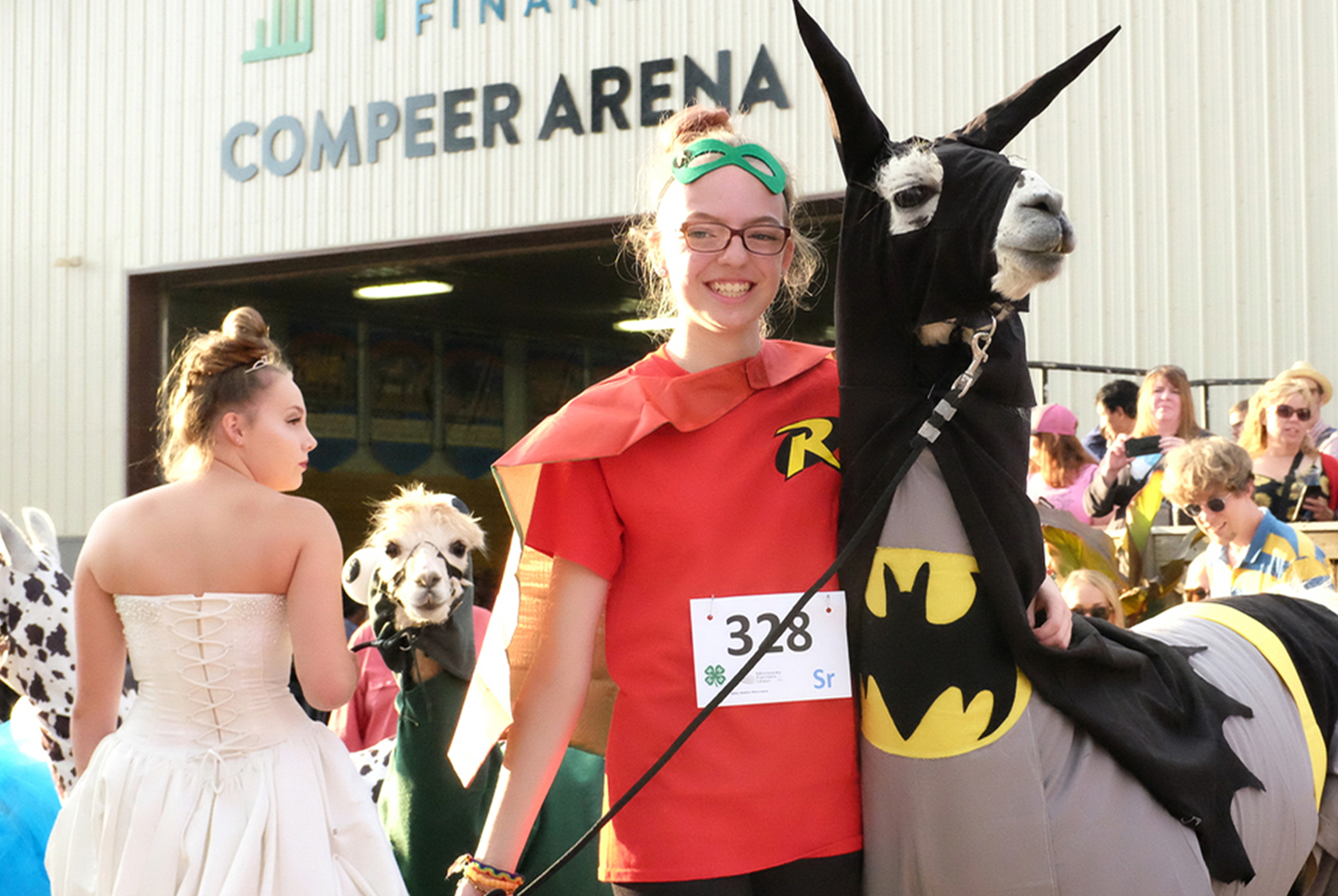 A teenage girl wears a Robin costume next to her llama dressed up as Batman