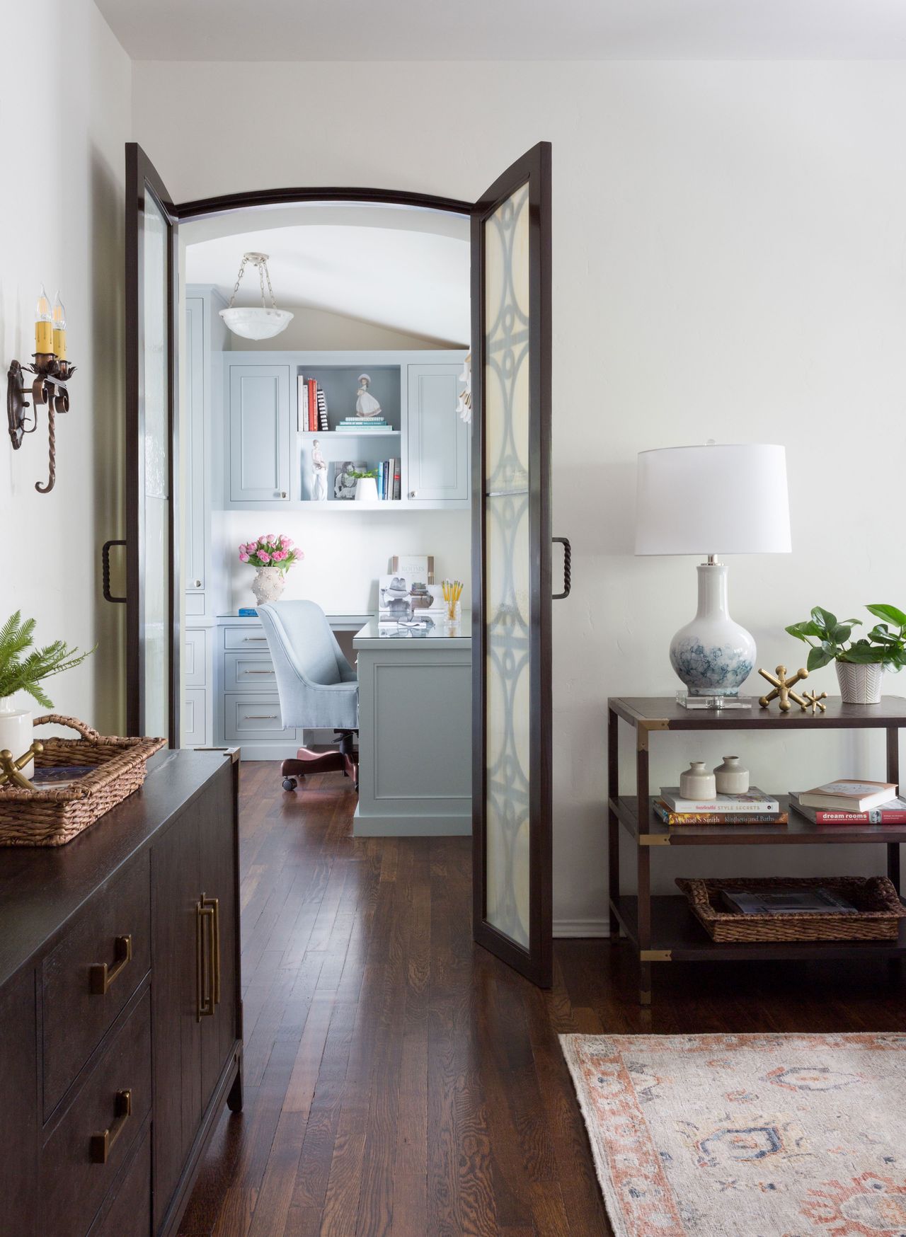 living room with white walls and dark wooden sideboard with double doors open onto home office with cupboards and desk chair
