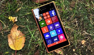 A smartphone with a colorful app tile interface lies on grass surrounded by autumn leaves. The bright screen contrasts with the earthy background.