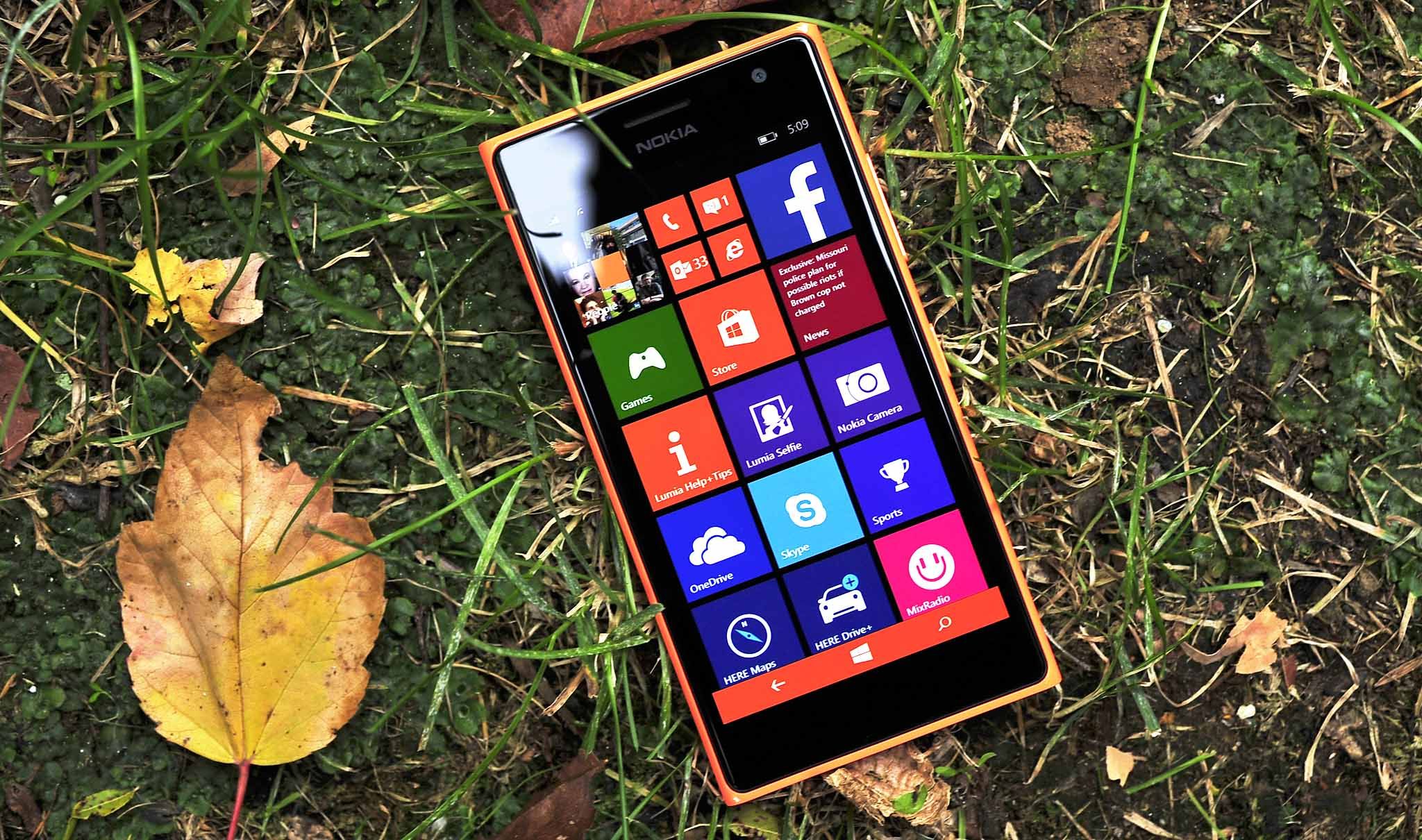 A smartphone with a colorful app tile interface lies on grass surrounded by autumn leaves. The bright screen contrasts with the earthy background.