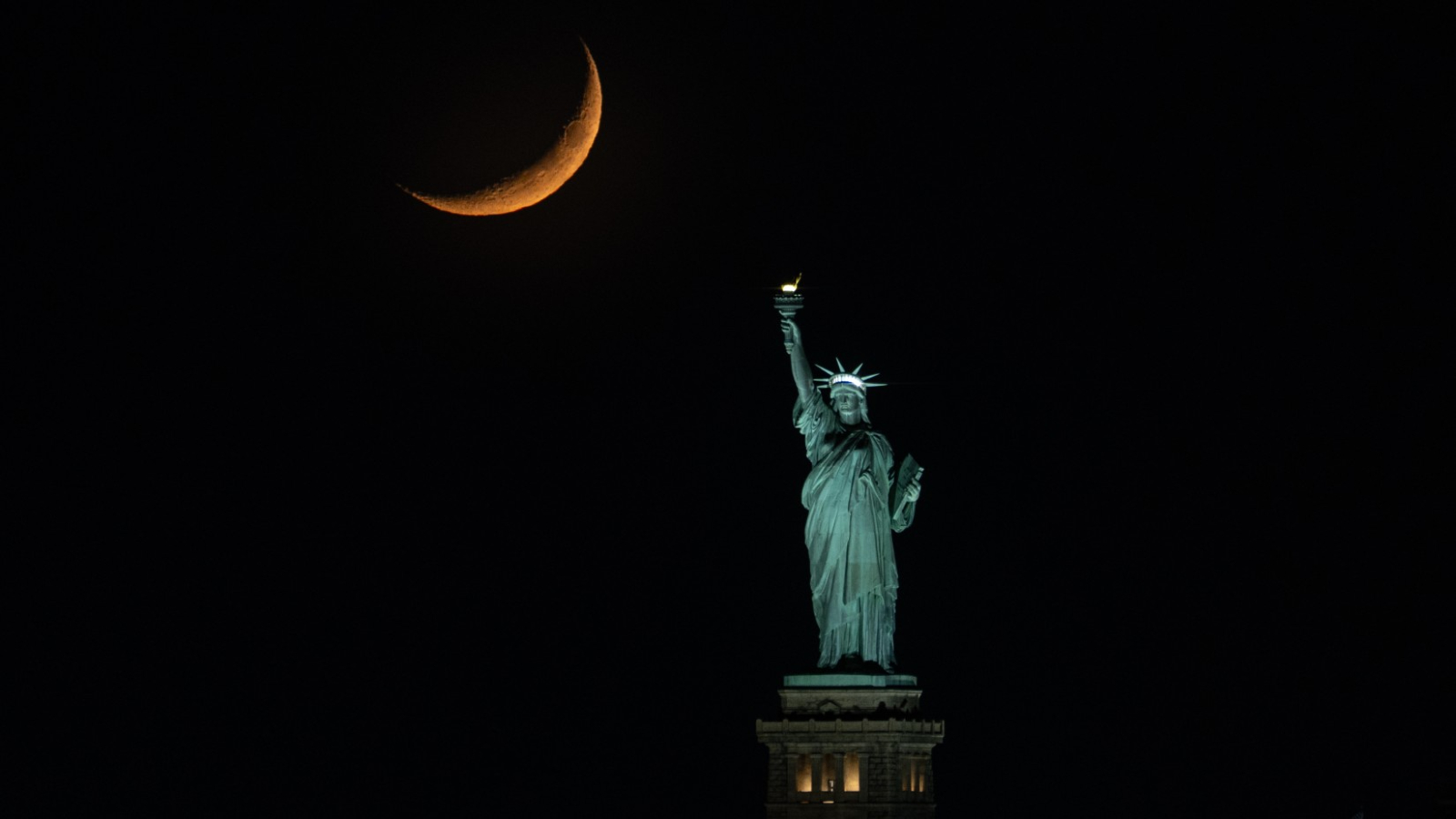 An orange crescent moon is pictured to the upper left of the Statue of Liberty in a dark sky.