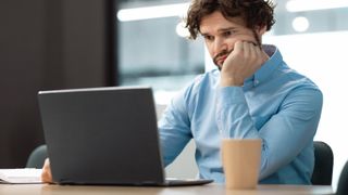 A man sitting at a table and looking at a laptop with a puzzled expression, holding his hear up with his hand