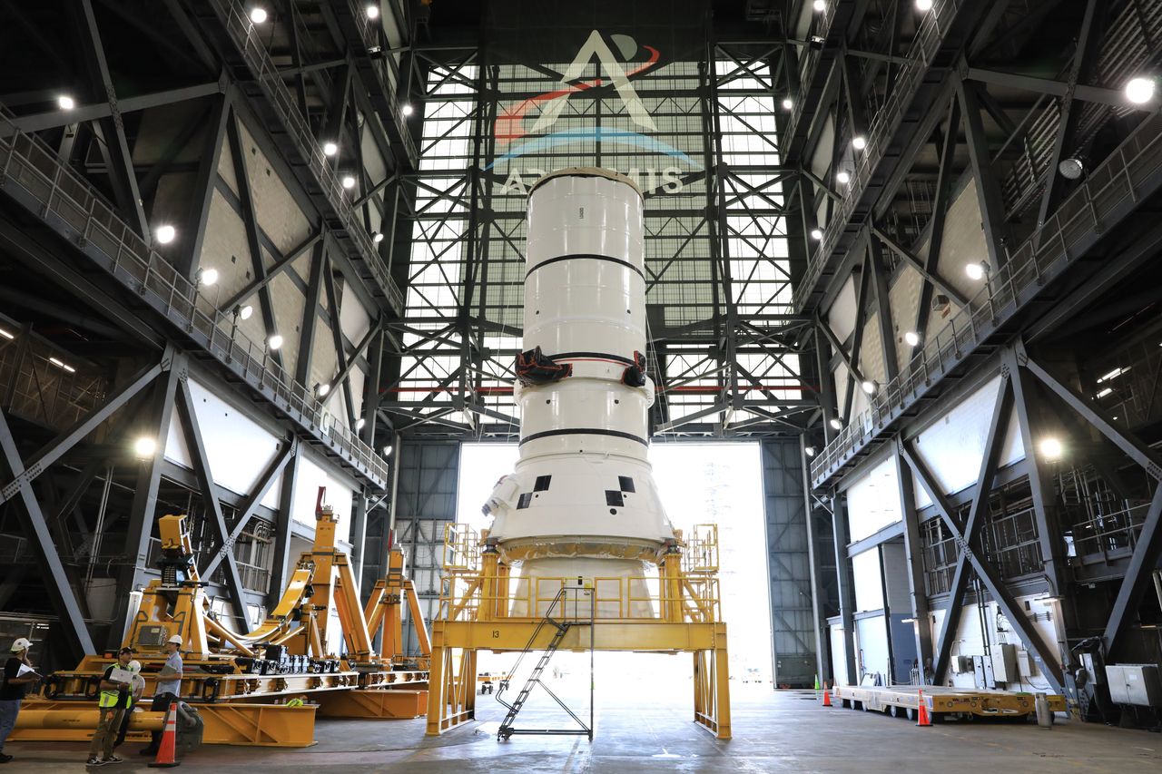 The bottom segment of a white solid rocket booster stands on a yellow support inside a giant rocket hangar.