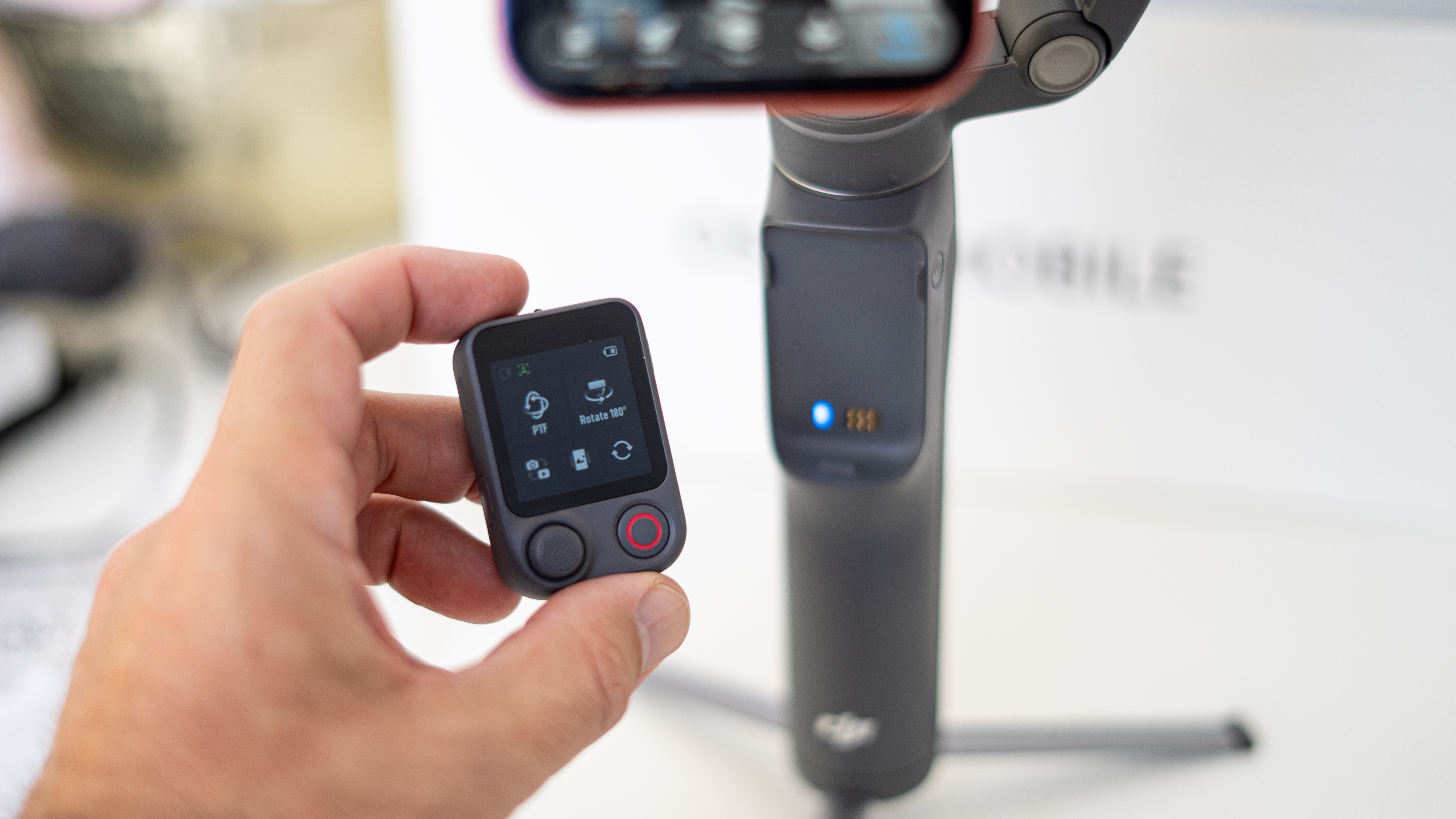 DJI Osmo Mobile 8P remote in Adam Juniper's hand. There is a connection light and metal contacts visible on the gimbal's handle behind.