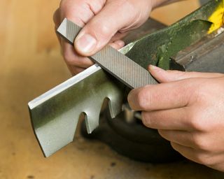 Man's hands are using a file to sharpen a lawnmower blade which is clamped in a vise atop a wood workbench. Focus is on the file near the bottom hand, the blade is slightly soft
