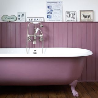 a pink freestanding clawfoot rolltop bathtub in front of a wall with matching colour panels, with a selection of bathroom signs on top of the panneling