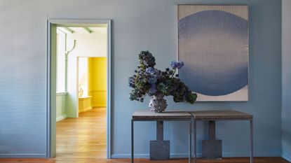 A blue-painted entryway with wooden flooring, a two-section console table with a vase of flowers and a blue artwork above it, plus a green room that leads onto a yellow room through the doorway