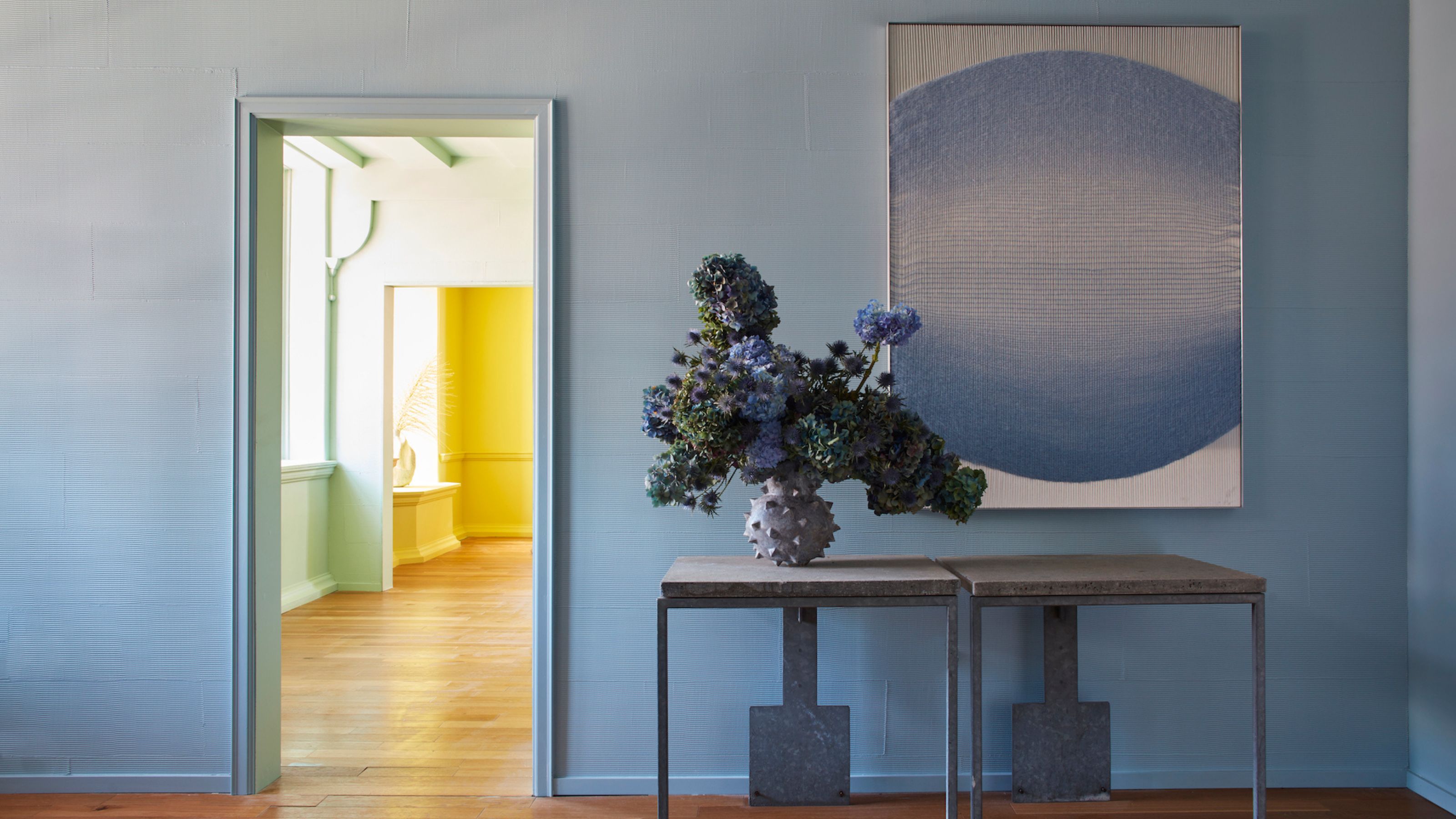 A blue-painted entryway with wooden flooring, a two-section console table with a vase of flowers and a blue artwork above it, plus a green room that leads onto a yellow room through the doorway