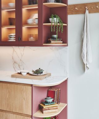 kitchen corner with burgundy glass-front cabinets, wooden open shelves, and a marble backsplash. Warm lighting highlights stacked dishes and plants, while a curved wooden countertop features a small display of books and cookware.