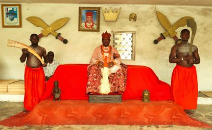 A portrait of an important tribal person sat on a red chair wearing a red/orange robe and a red crown. He is surrounded by two tribal persons holding wood-style objects. On the wall behind are portraits and large wood swords. 