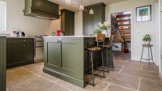 olive green Shaker kitchen with kitchen island with wooden bar stool and stone flooring