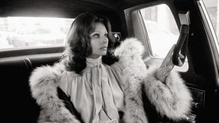 Sophia Loren is seen in a profile view sitting in the back of a car, wearing a voluminous fur-trimmed coat and a pussy-bow blouse.