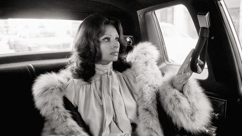 Sophia Loren is seen in a profile view sitting in the back of a car, wearing a voluminous fur-trimmed coat and a pussy-bow blouse.