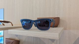 Une paire bleue de lunettes intelligentes Ray-Ban Meta Smart Glasses Collection sur une table en bois, devant leur boîtier de chargement.