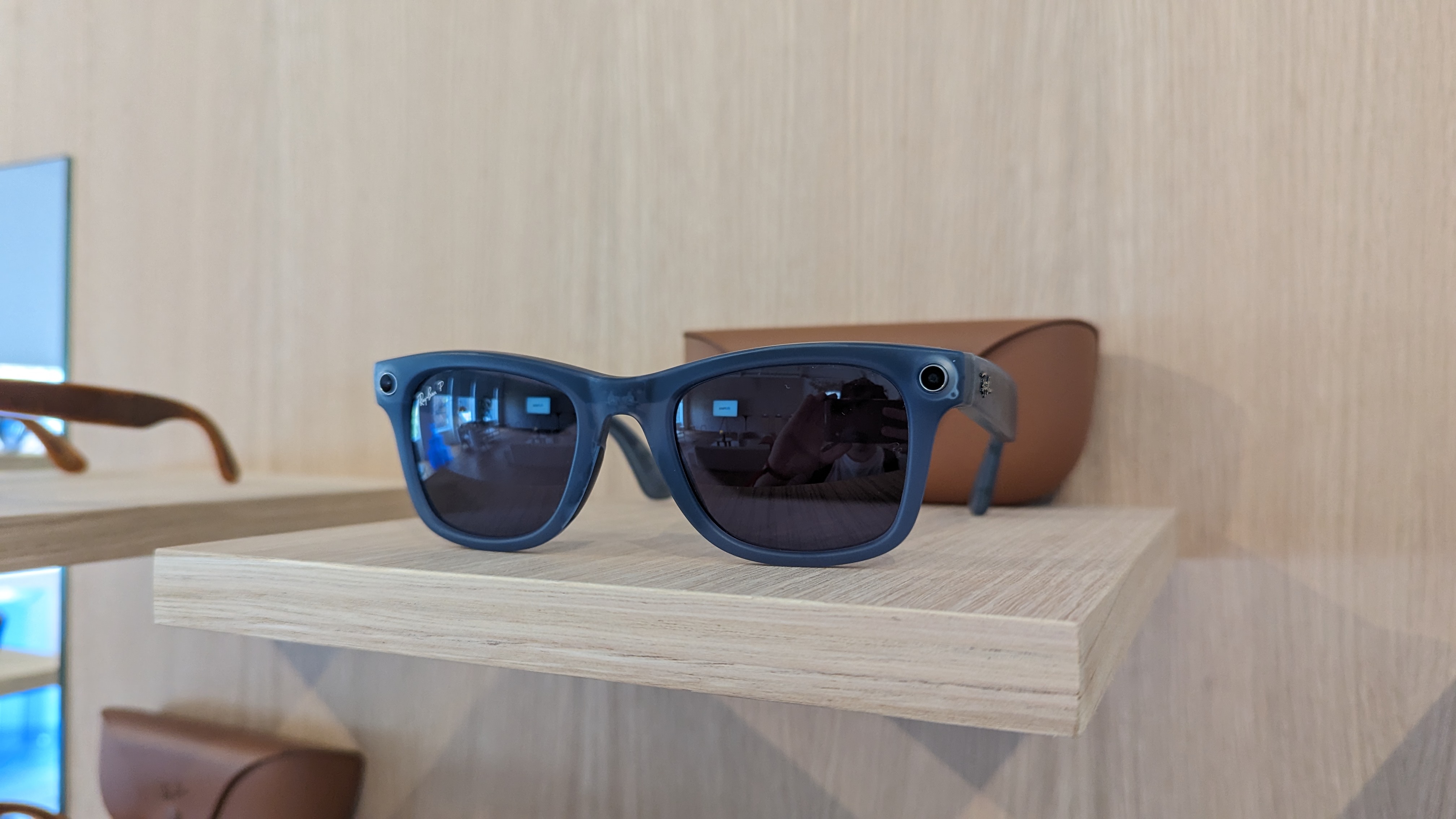 A blue Wayfarer pair of the Ray-Ban Meta Smart Glasses Collection on a wooden table in front of their charging case