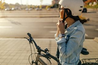 A woman buckles her bike helmet while standing over bike - a side-on shot.