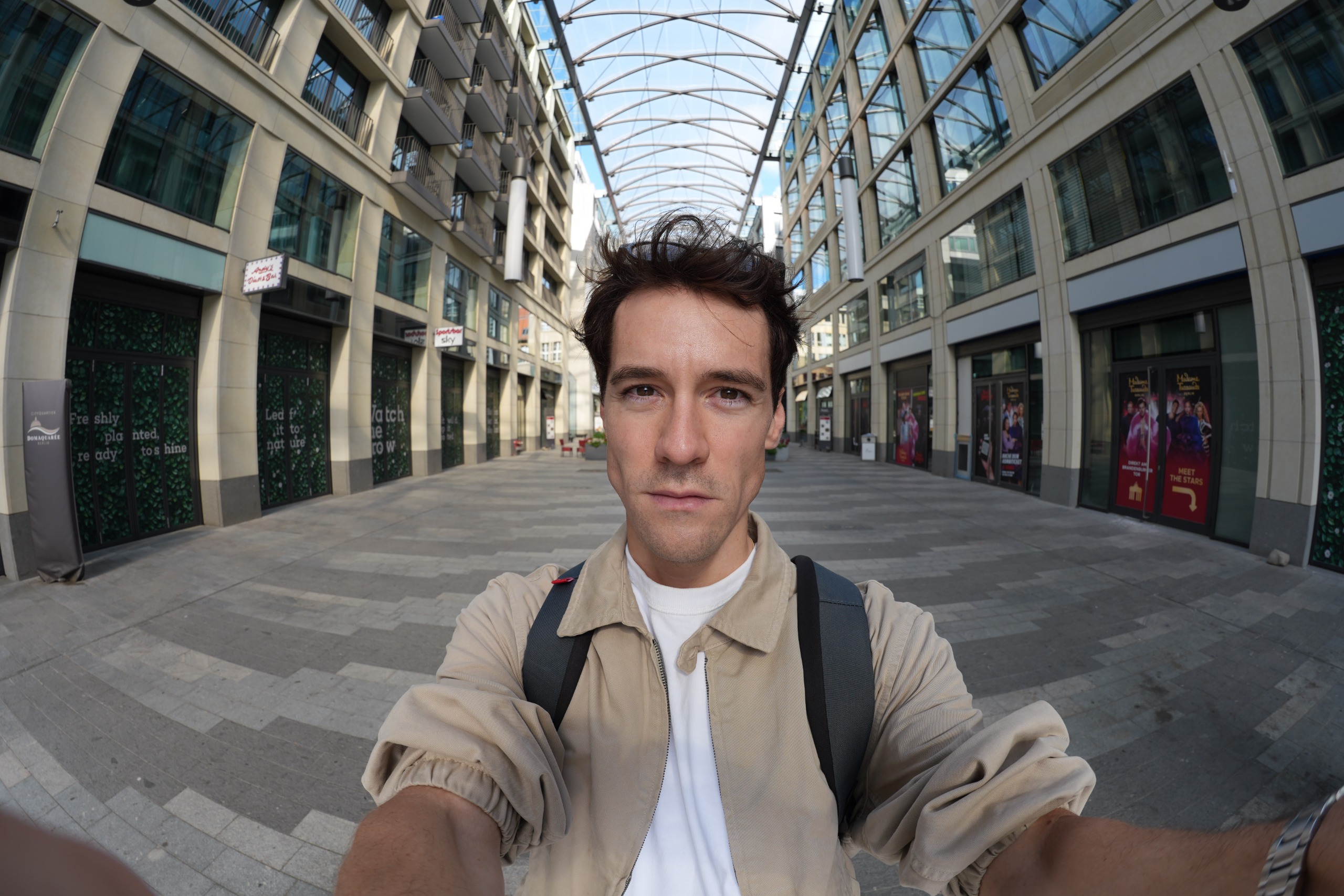 Wide-angle selfie of a man standing in a modern covered pedestrian courtyard in Berlin.