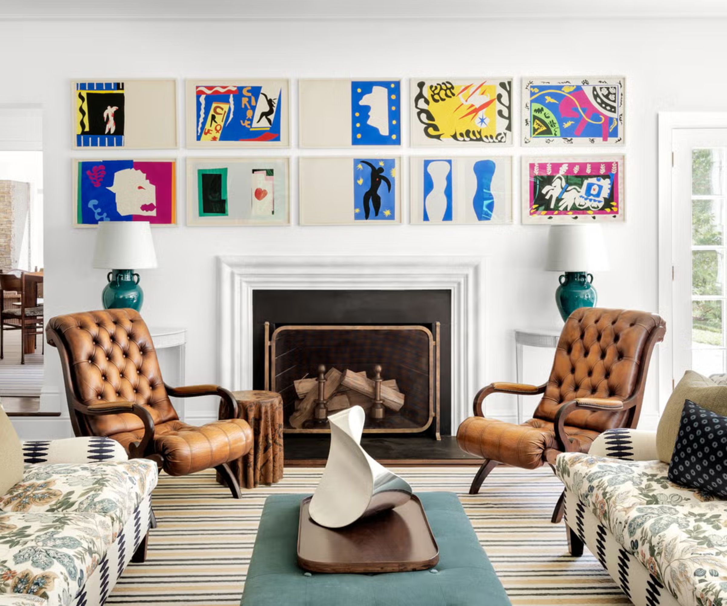 White living room with fireplace with brightly colored artwork hungover. Two patterned sofas and two brown leather armchairs sit around the fireplace on top a striped rug