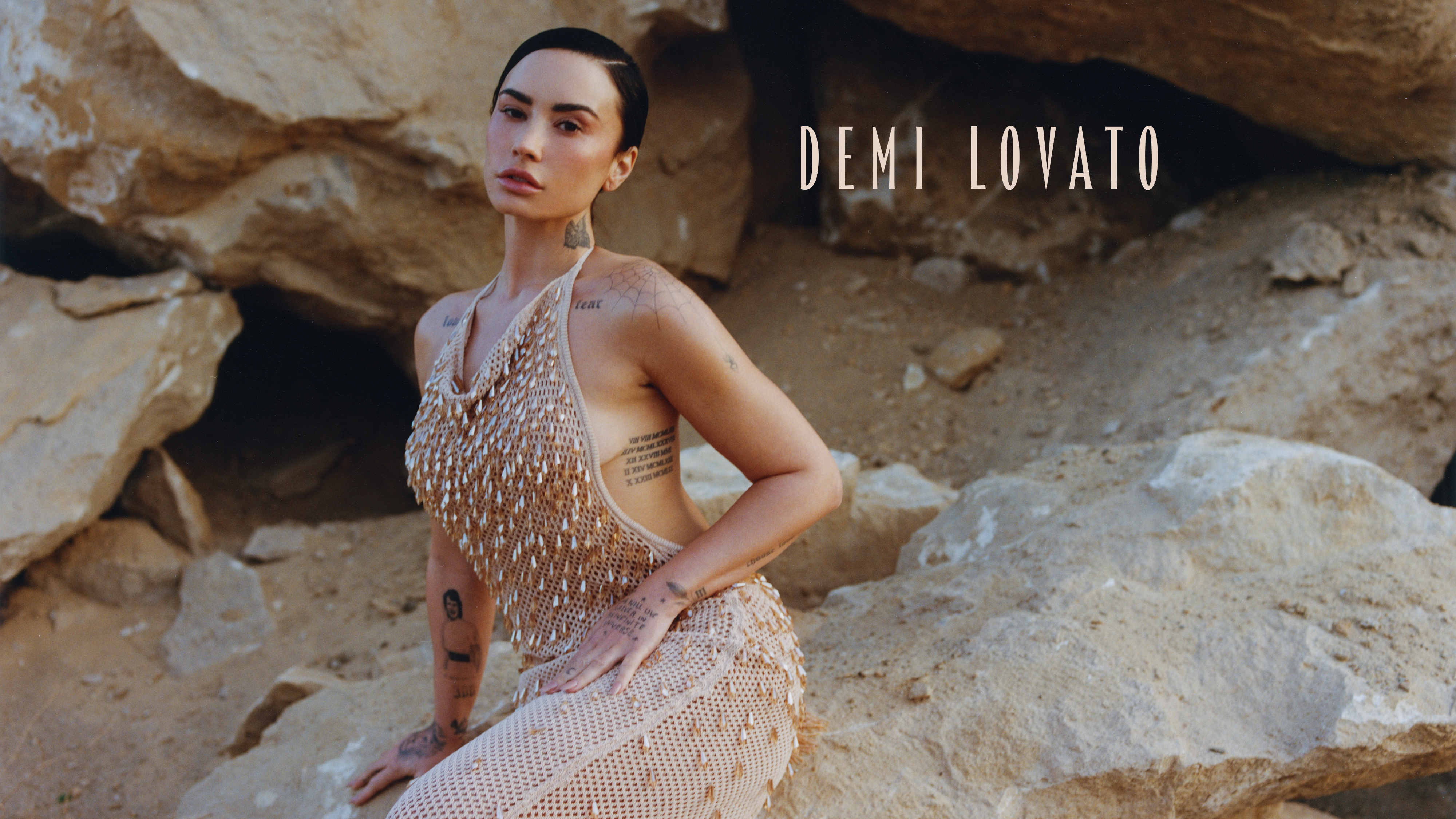 Demi Lovato sits surrounded by rocks. She is wearing a nude dress made from a netted material and embellished with crystals. 