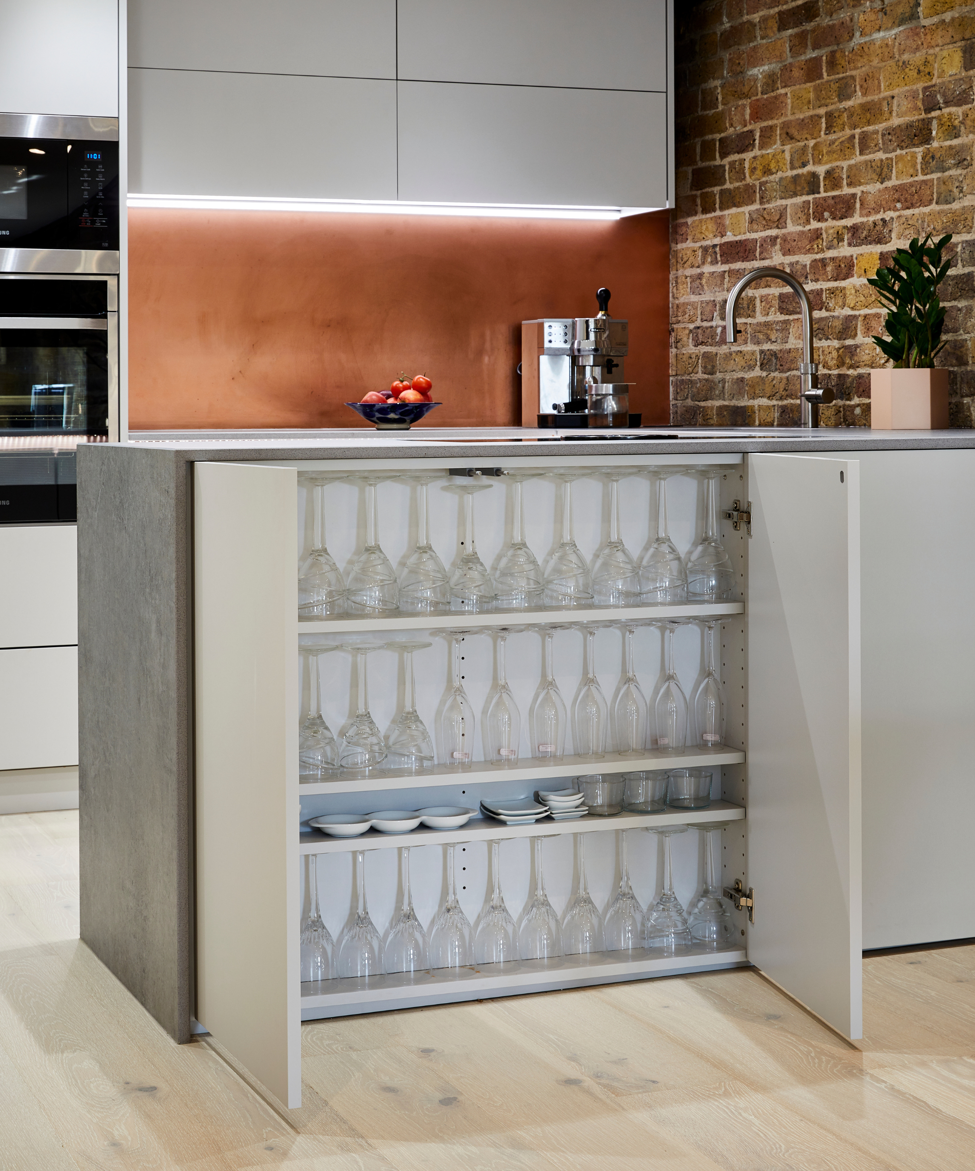 A concealed cupboard opens to reveal neatly arranged rows of glasses and plates within a modern kitchen island. Brick walls, copper splashback and sleek cabinetry create a stylish backdrop, blending practical hidden storage with contemporary design.