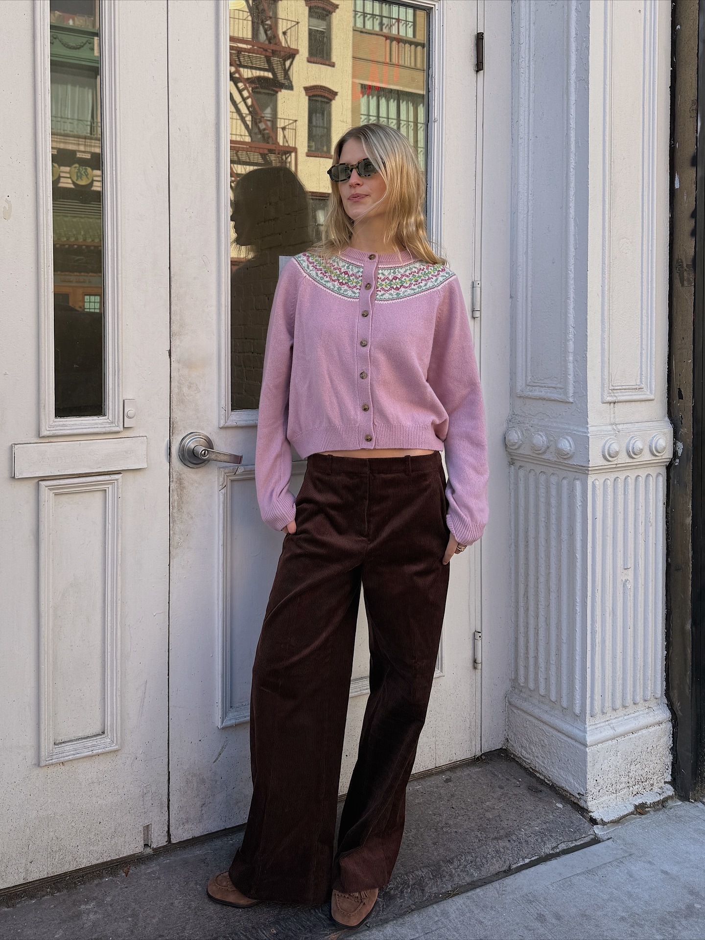 An image of an influencer wearing a pink cardigan, brown relaxed trousers and brown loaders in New York City