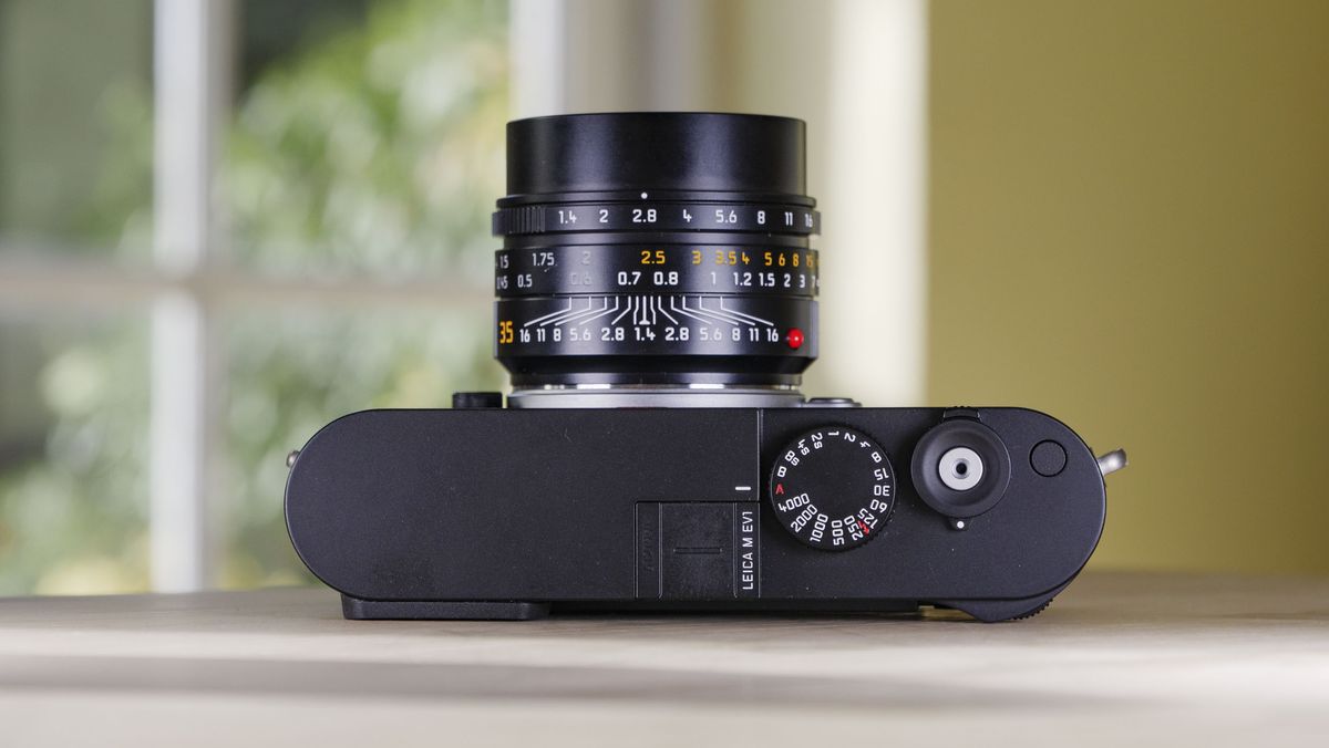 Top plate of the Leica M EV1 camera, which is resting on a wooden table with glass-panelled door backdrop