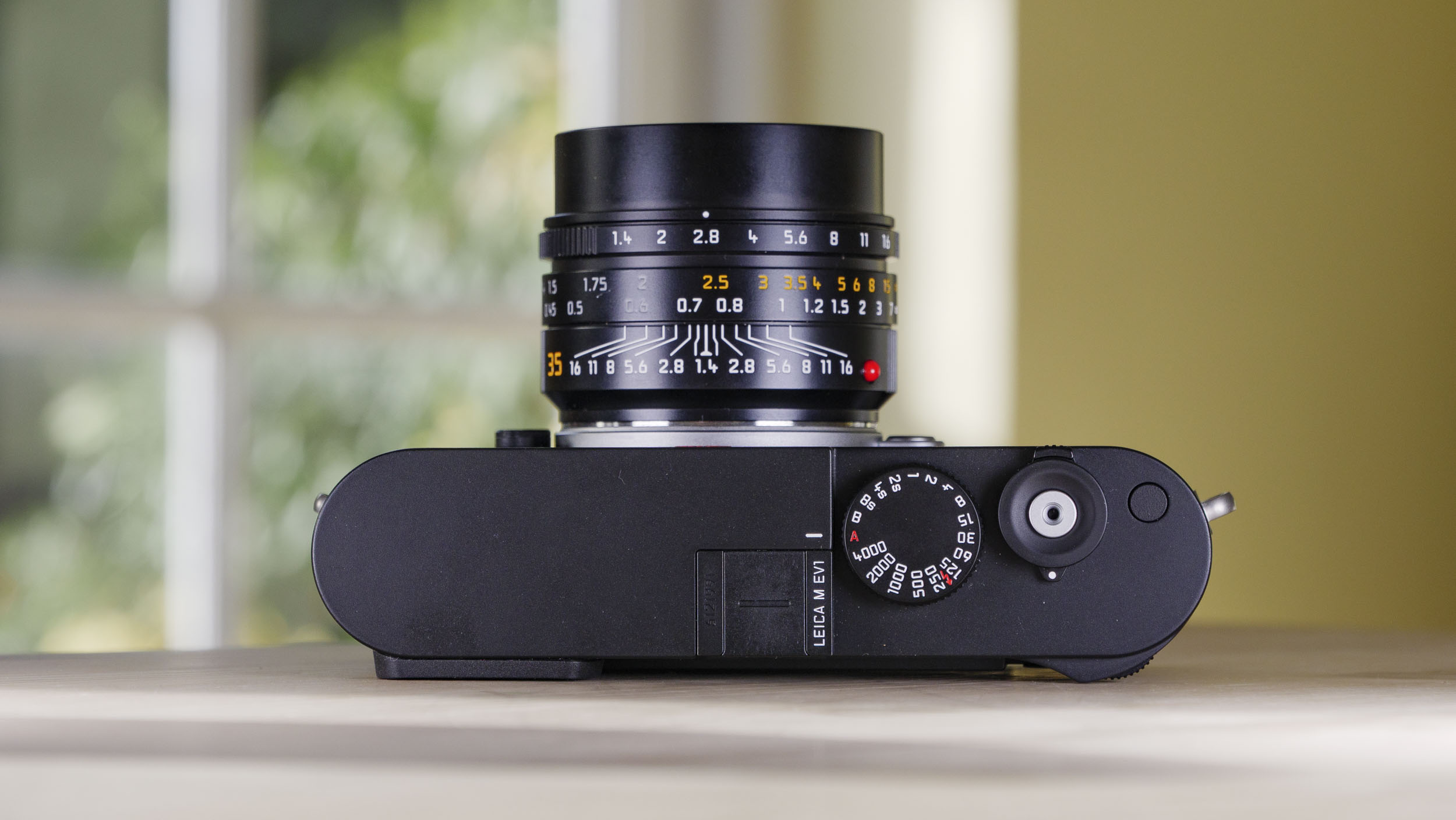 Top plate of the Leica M EV1 camera, which is resting on a wooden table with glass-panelled door backdrop