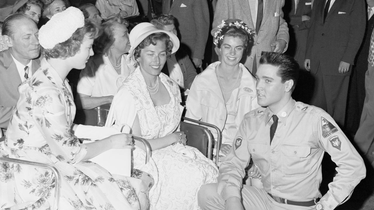 A black-and-white photo of Elvis Presley wearing a military uniform kneeling in front of Princesses Margrethe of Denmark, Astrid of Norway, and Margaretha of Sweden.