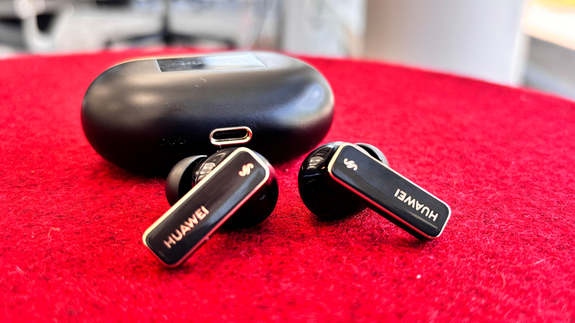 Huawei FreeBuds Pro 4 in-ear headphones on red fabric surface in front of charging case