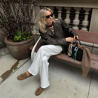 Lucy Williams Wearing Brown Leather Jacket and White Pants Sitting on Bench