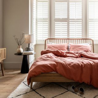 A wooden bedframe in front of a large bay window with white shutters. The bed is dressed with terracotta linen bedding