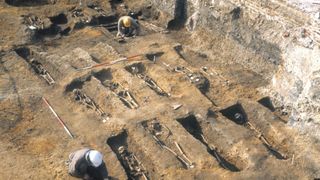 a historic photo of the plague pits in london being excavated; archaeologists can be seen examining skeletal remains in a line of open graves