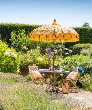 A garden with a yellow parasol and matching cushions resting on wooden chairs