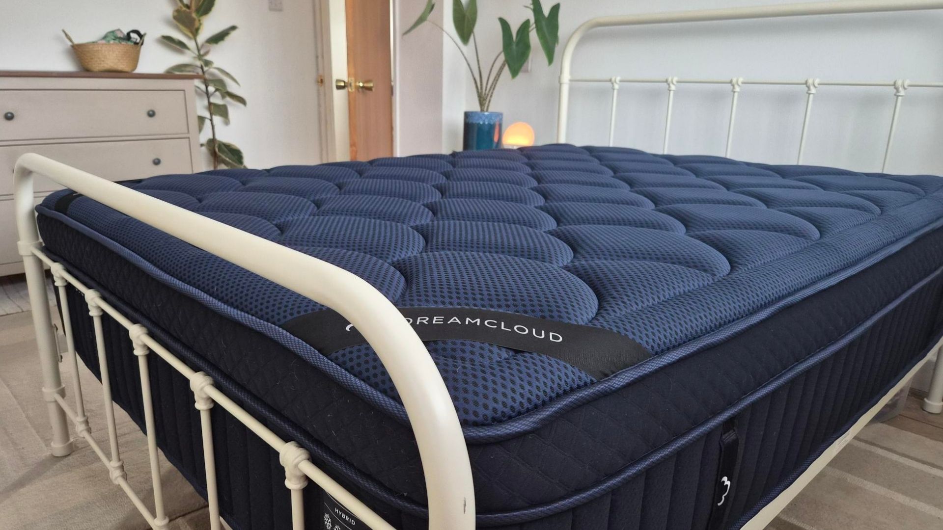 The image shows a navy blue mattress on a white bedframe, placed within a neutrally decorated bedroom.