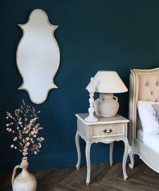 A blue bedroom with a mirror, table lamp, and beige bed