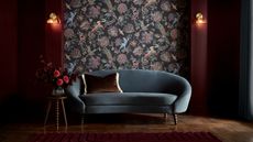 Image of a dark entryway painted in a deep burgundy paint color with a mural on the center wall. The mural is black with multicolored patterns. There is a curved blue velvet sofa in the room.