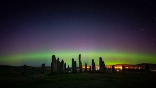stone pillars stand up in shadow against the dark horizon. the rim of the northern lights is visible in behind, along with a hill and city lights. stars are in the sky above